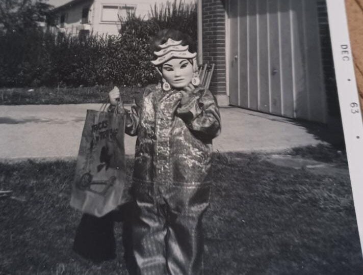 A child in a shiny costume and a mask stands outside by a garage, holding a decorated trick-or-treat bag. The photo is black and white and dated December 1963.