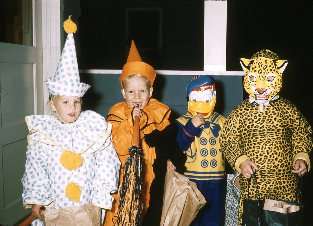 Four children in vintage Halloween costumes stand side by side holding treat bags. Their costumes include a clown, a wizard or jester, Donald Duck, and a leopard. They are smiling or posing near a doorway.
