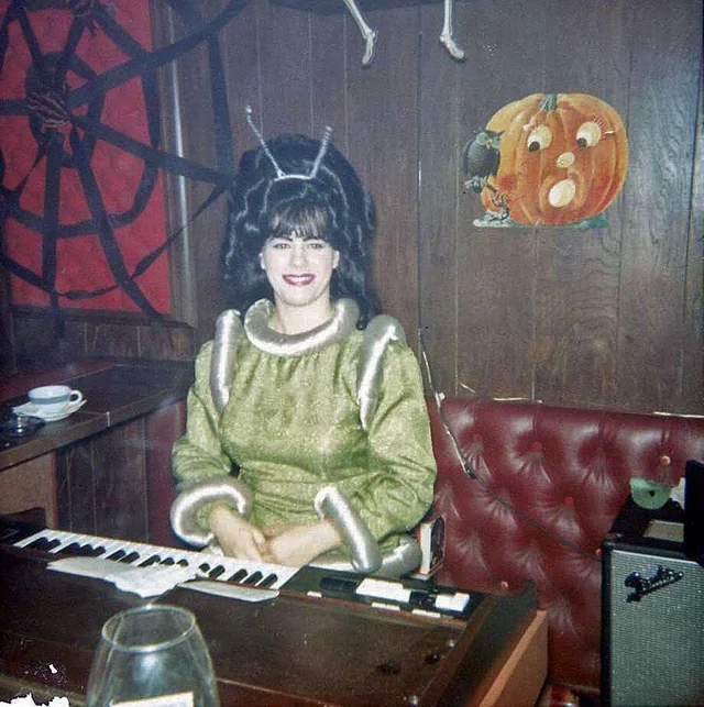 A woman in a green bug-themed costume with antennae sits at a keyboard in a wood-paneled room decorated with a large spiderweb and a pumpkin drawing. She is smiling and seated on a red cushioned bench.
