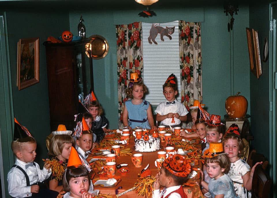 A group of children wearing party hats sit around a table decorated for Halloween with festive tableware, pumpkin decorations, and a cake. The room has green walls and floral curtains, with Halloween decor throughout.