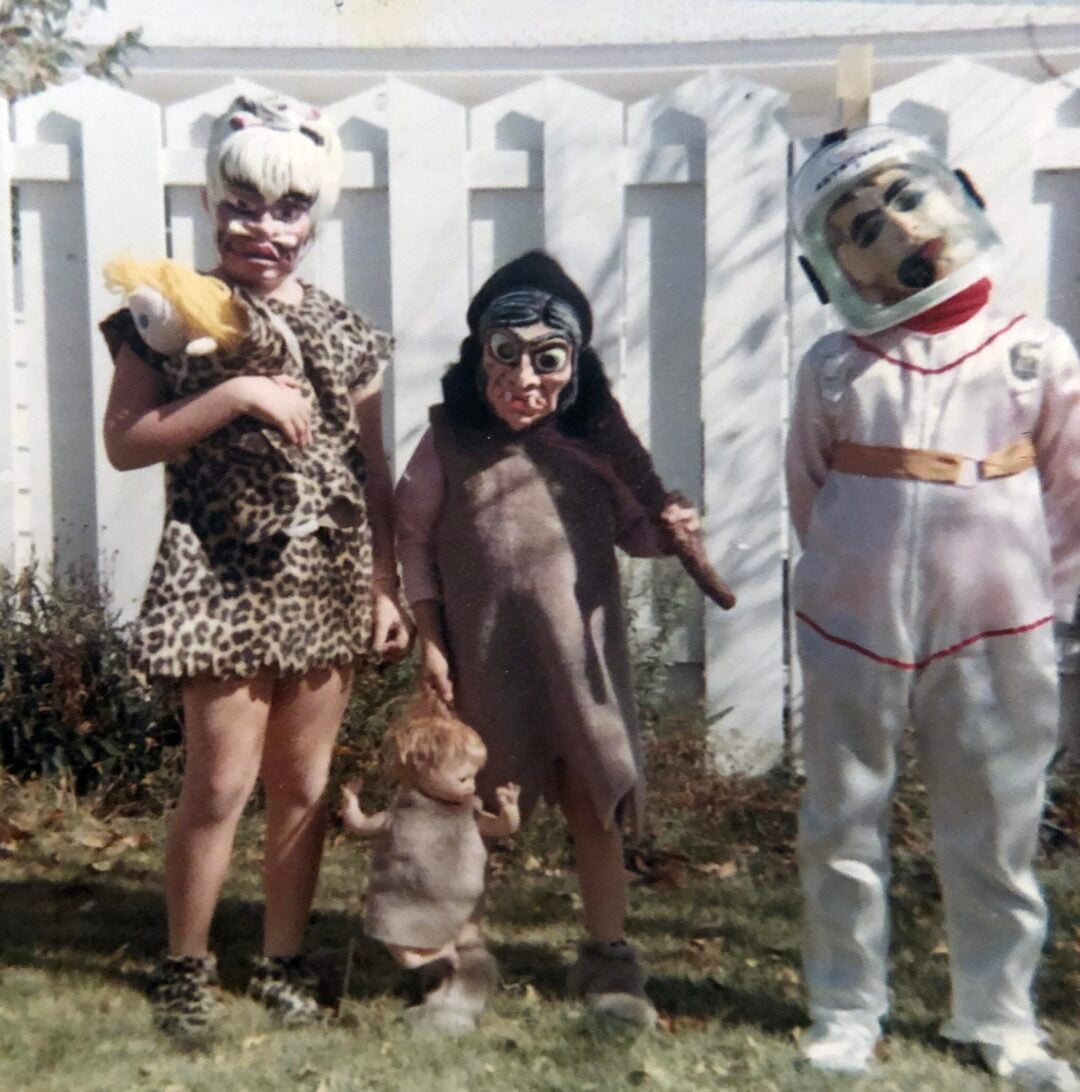 Three children in vintage costumes stand in front of a white picket fence. One is dressed as a cavewoman, another as a caveman, and the third as an astronaut. A toddler in a brown outfit stands in front of them.