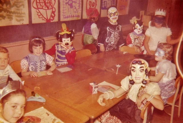 A group of young children in colorful costumes and masks sit and stand around a classroom table, with artwork displayed on the wall behind them. Some children appear to be coloring or drawing.