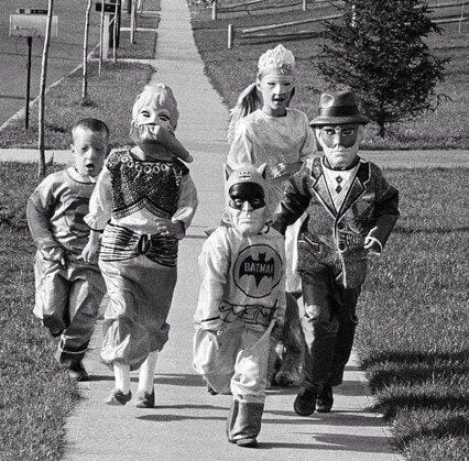 Five children in vintage costumes, including Batman and a clown, walk excitedly down a suburban sidewalk on a sunny day, likely trick-or-treating in a residential neighborhood.