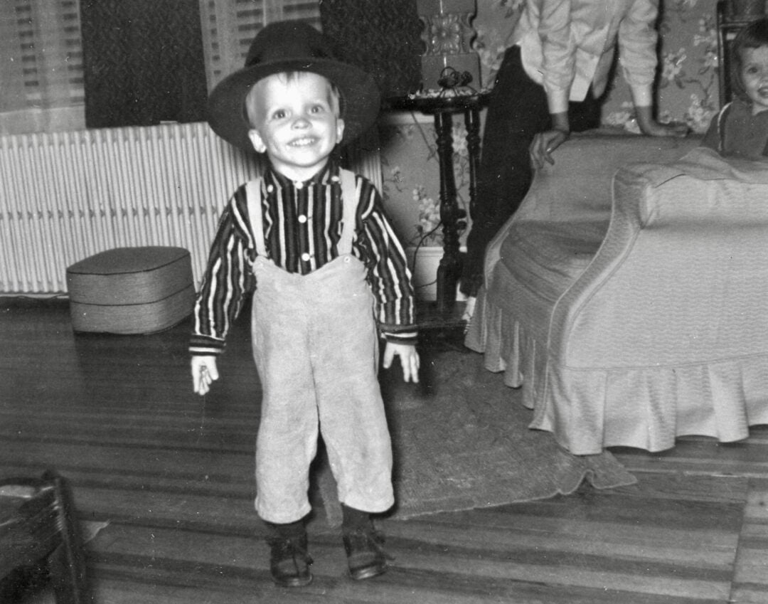 A young child wearing a large hat, striped shirt, and suspenders stands and smiles in a living room with wooden floors and patterned wallpaper. An adult is partly visible behind a sofa in the background.