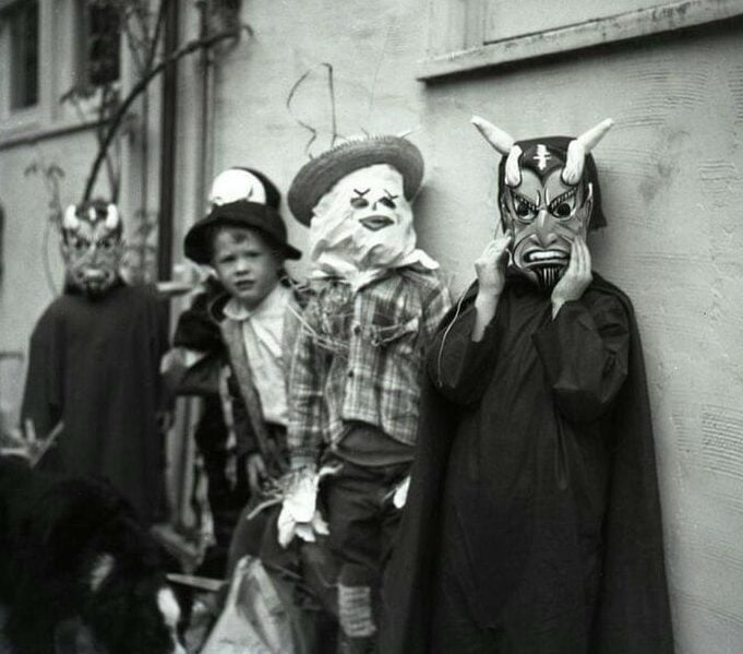 Four children stand in costume for Halloween; two wear scary masks, one is dressed as a cowboy or scarecrow, and another wears a painted clown face. They stand outside against a building, looking toward the camera.