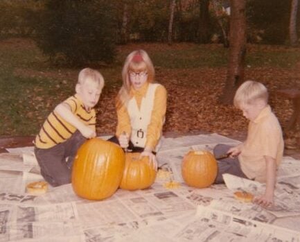 Three children sit on newspapers outdoors, carving pumpkins together on a fall day. Leaves cover the ground and trees in the background, suggesting an autumn setting.