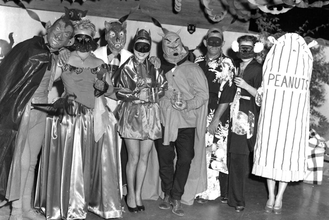 A group of people in elaborate masks and costumes, including animal masks, capes, and one person dressed as a peanut, stand together at a party with festive decorations overhead.