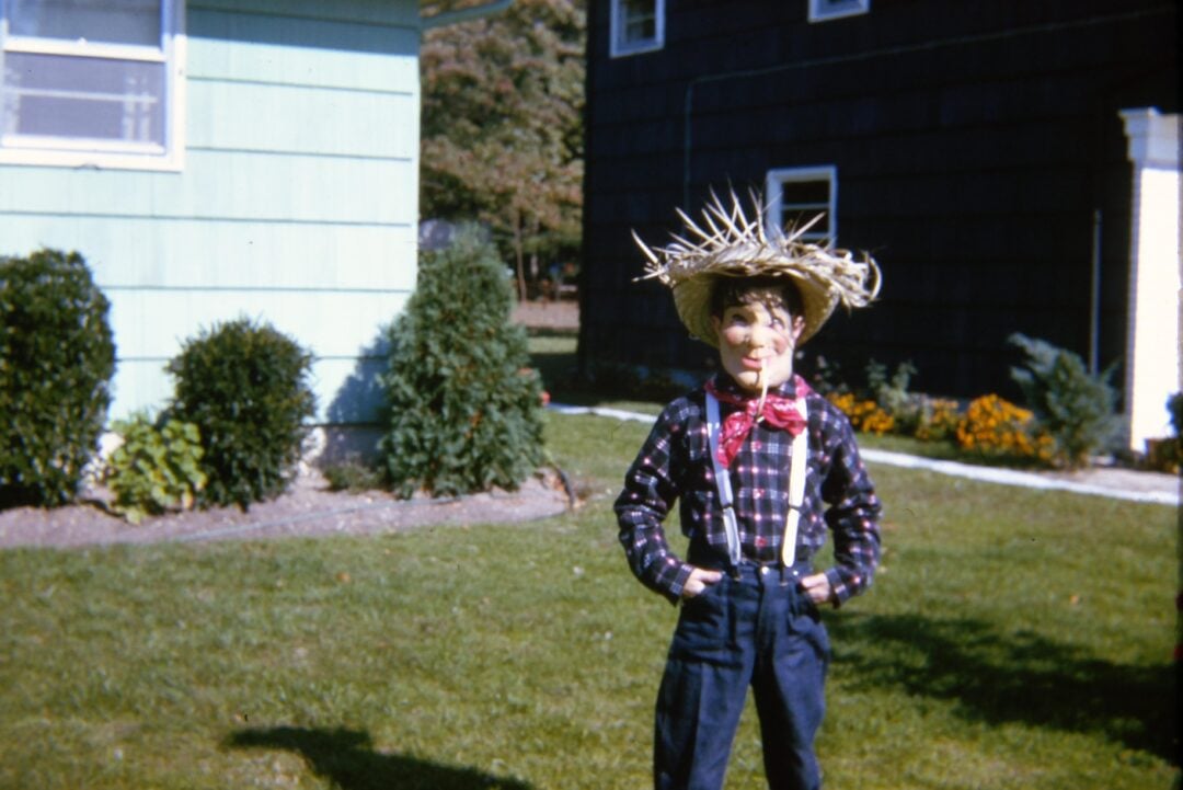 A child dressed as a scarecrow, wearing a straw hat, plaid shirt, red bandana, suspenders, and straw details, stands on a lawn in front of two houses with bushes and flowers in the background.