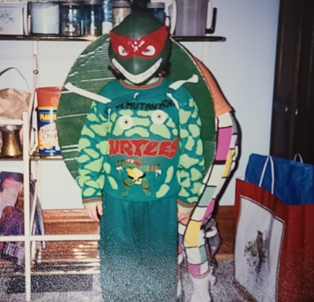 A child dressed in a Teenage Mutant Ninja Turtles costume, featuring a green mask, a sweater with the turtle design, and a shell accessory, stands indoors near shelves and gift bags.