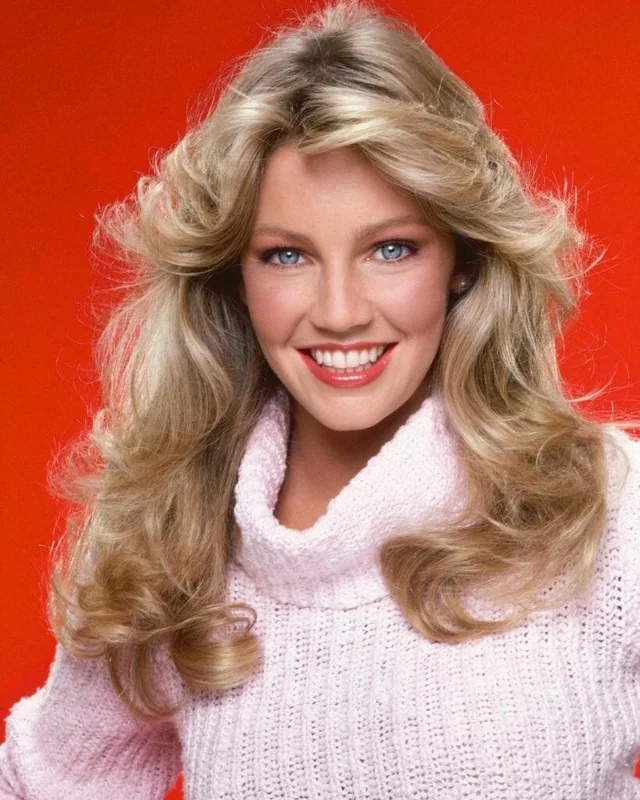 A woman with long, wavy blonde hair and blue eyes smiles at the camera. She is wearing a light pink turtleneck sweater. The background is bright red.