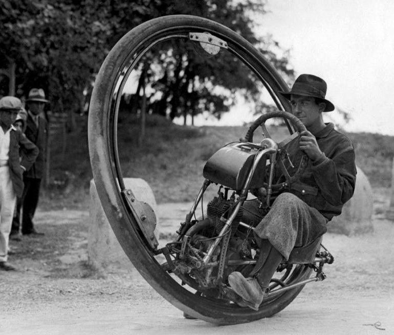 A man wearing a hat and coat rides a large, single-wheeled motorized vehicle, known as a monowheel, on a dirt road while several people watch in the background. The image is in black and white.