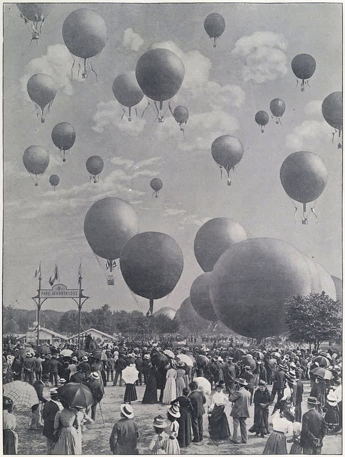A black-and-white vintage photo of a large crowd gathered outdoors, watching numerous hot air balloons rise into the sky during a balloon festival or event.