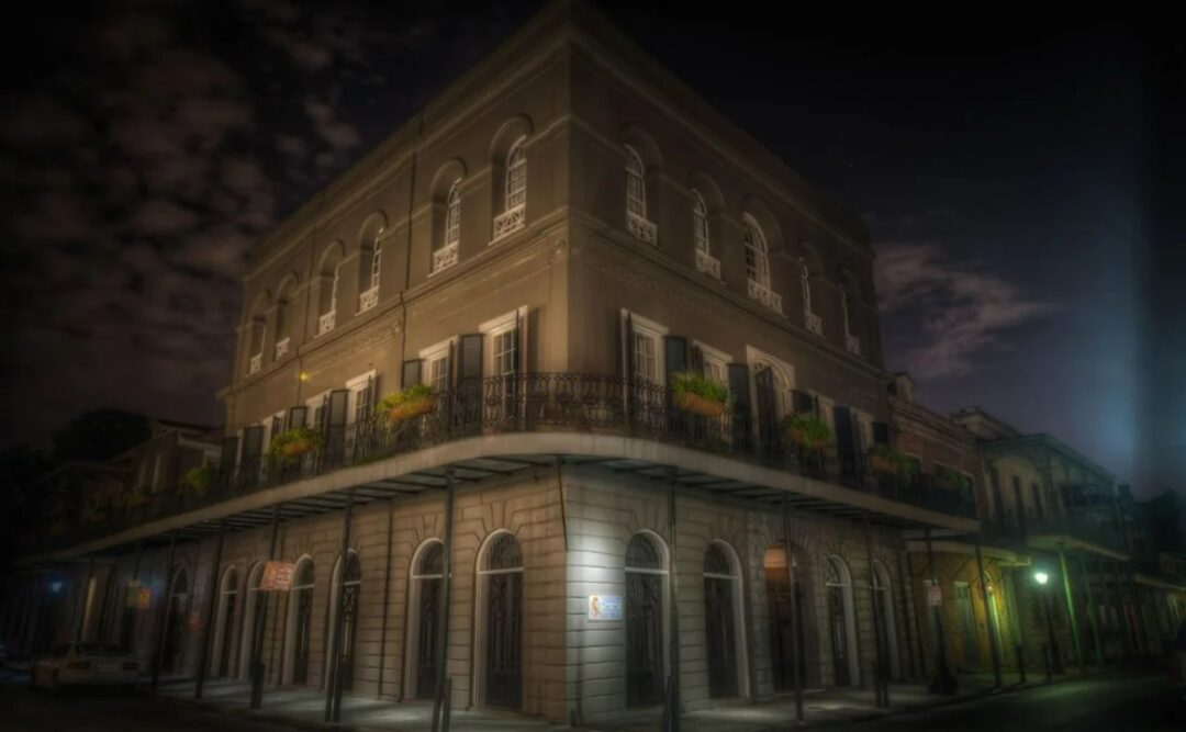 A historic, three-story building with arched windows, iron balconies, and potted plants sits on a street corner at night, illuminated by streetlights under a cloudy sky.