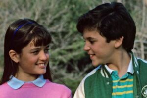 A young girl in a pink sweater smiles at a boy in a green jacket with a Jets logo. They are standing outdoors with blurred trees in the background, looking at each other warmly.