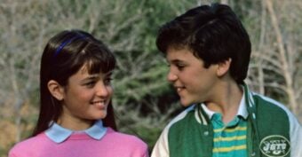A young girl in a pink sweater smiles at a boy in a green jacket with a Jets logo. They are standing outdoors with blurred trees in the background, looking at each other warmly.
