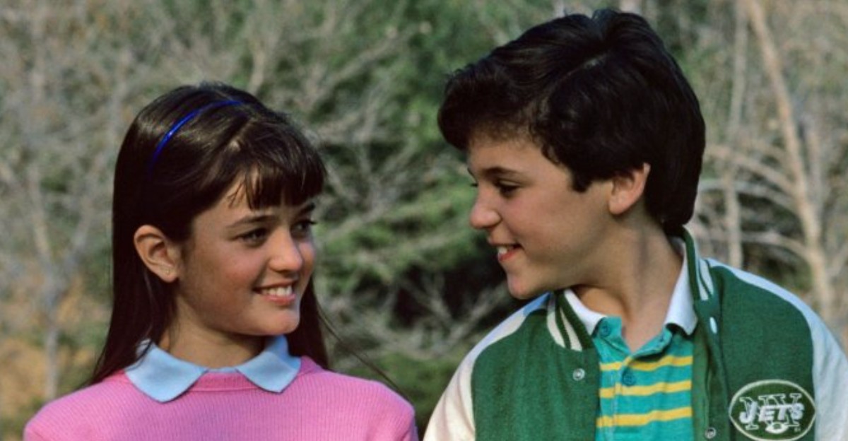 A young girl in a pink sweater smiles at a boy in a green jacket with a Jets logo. They are standing outdoors with blurred trees in the background, looking at each other warmly.