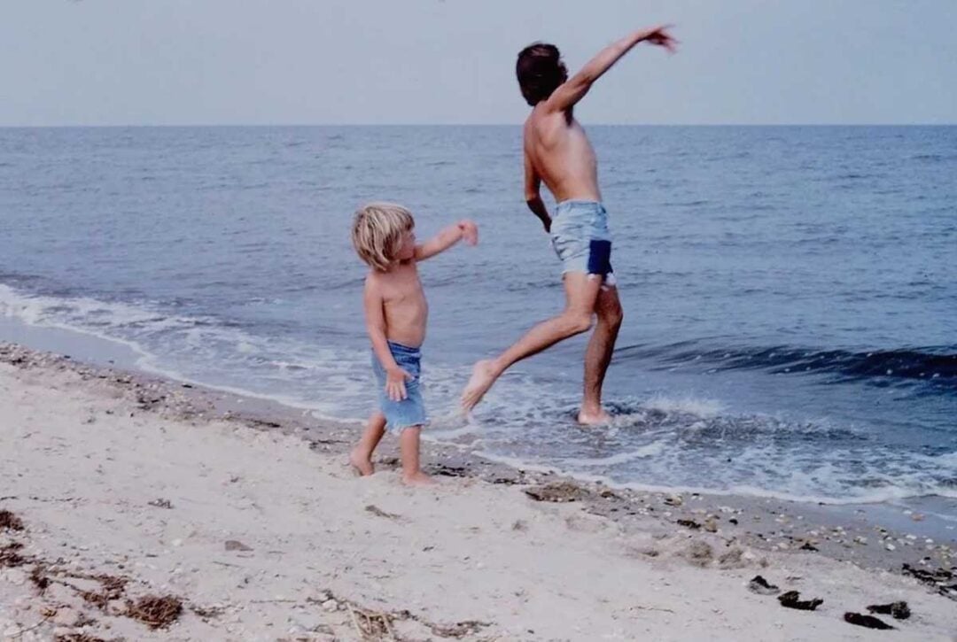 Two children in swimsuits play on a sandy beach near the water; one is throwing something into the sea while the other watches. The sky and ocean are calm in the background.