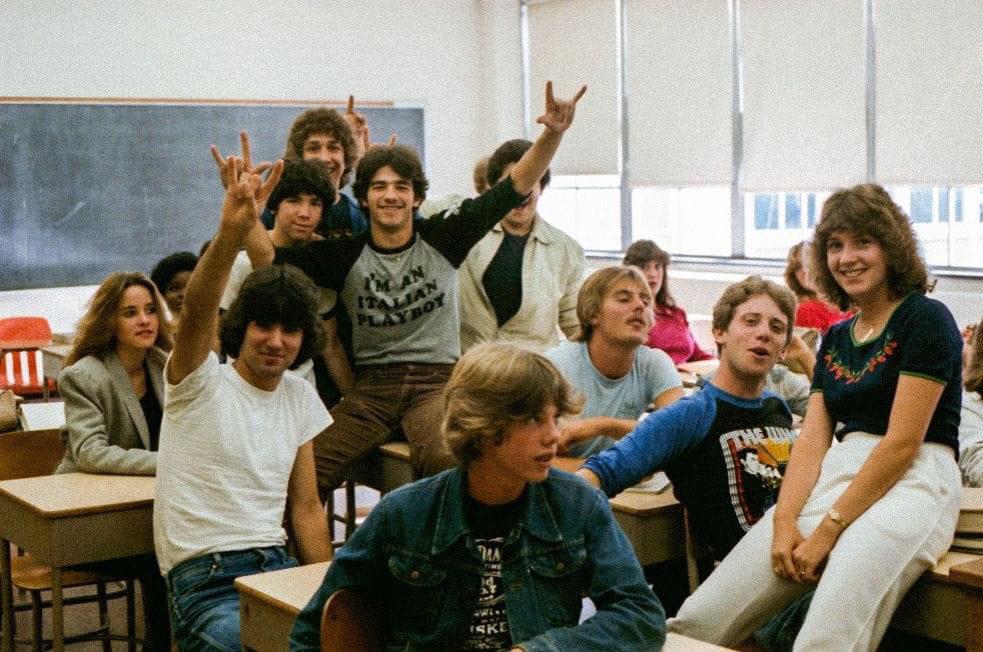 A group of teenagers pose and smile together in a classroom, sitting and standing around desks. Some make playful hand signs. The atmosphere is relaxed and casual.