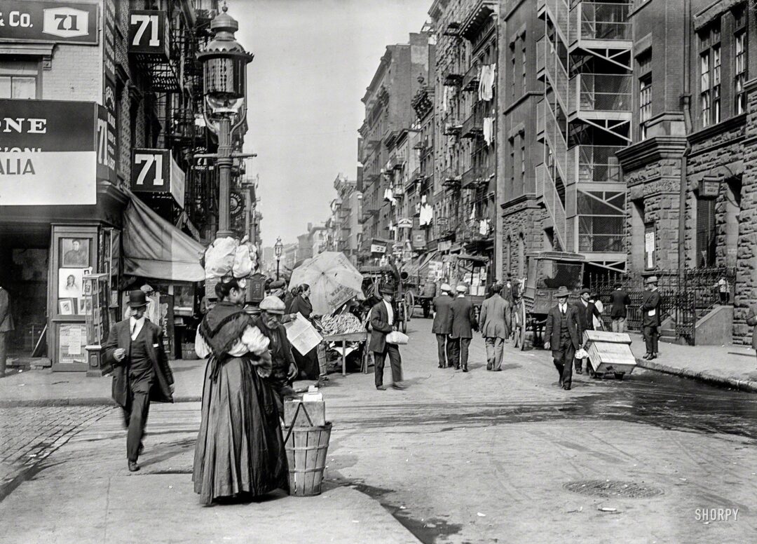 Black-and-white photo of a busy city street in the early 1900s, with people walking, street vendors selling goods, horse-drawn carts, and laundry hanging from tenement buildings.