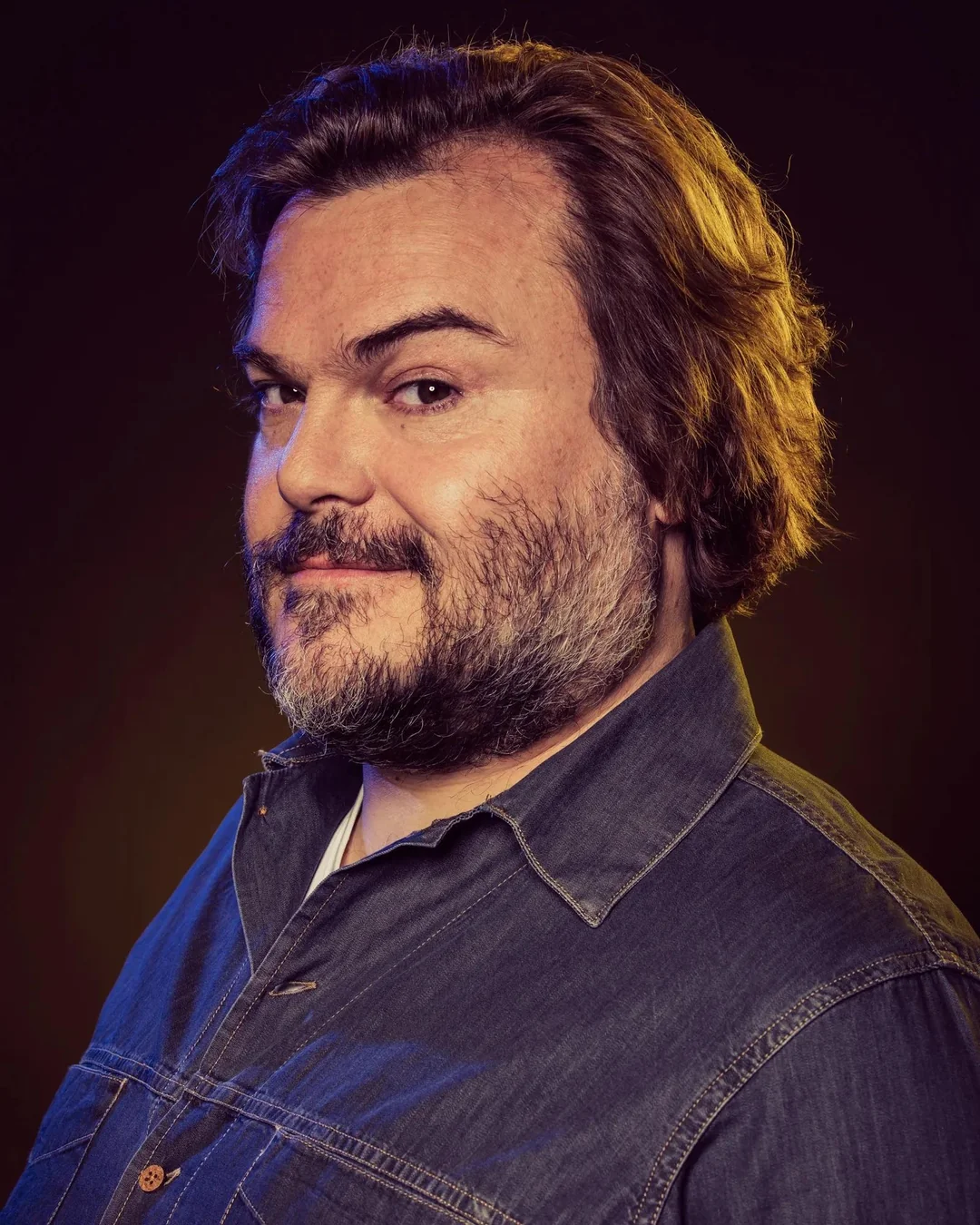 A man with medium-length brown hair and a beard is wearing a denim shirt and looking at the camera with a slight smile. The background is dark with warm, subtle lighting highlighting his face and hair.