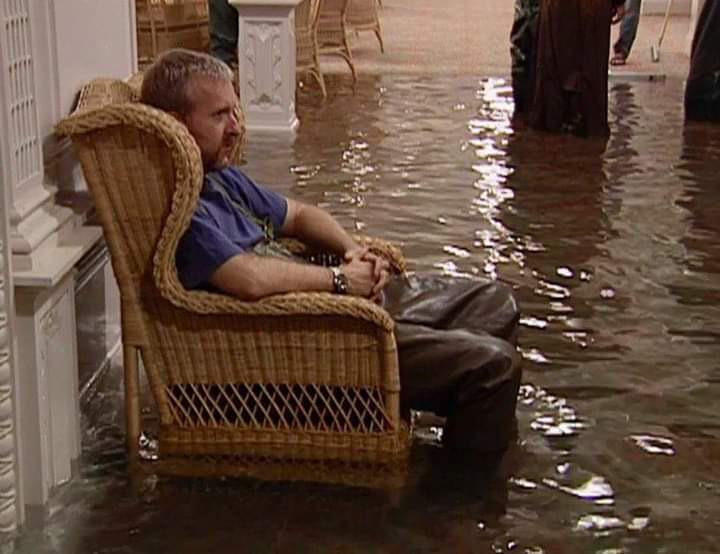 A man sits calmly in a wicker chair with his feet submerged in several inches of floodwater inside a building. The water covers the floor, reflecting tables and chairs in the background.