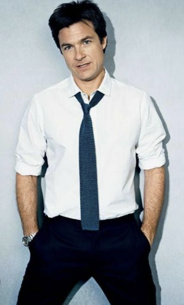 A man with short dark hair wearing a white dress shirt, loosened dark tie, and black pants stands against a light background with his hands in his pockets, looking at the camera with a slight smile.