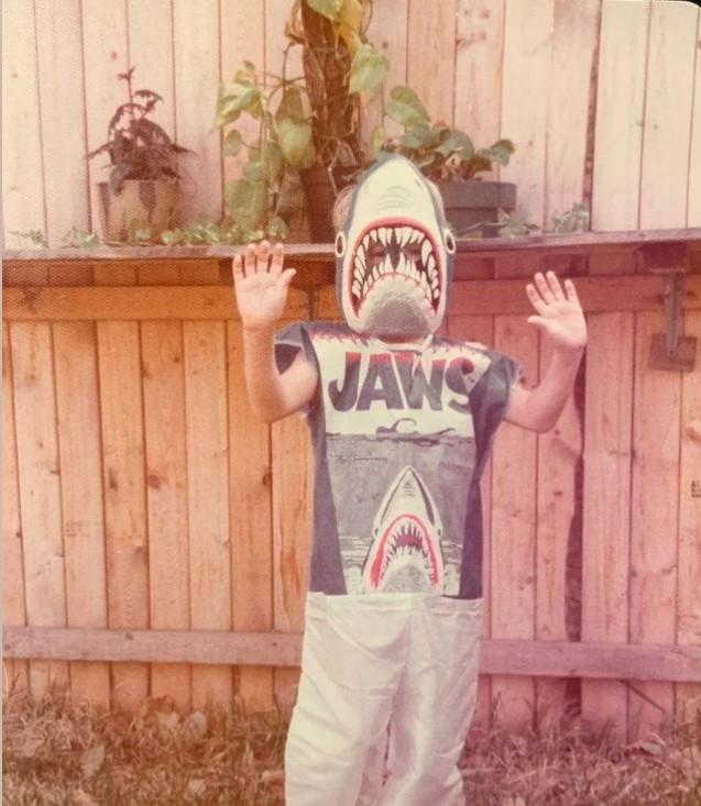 A child stands outdoors in front of a wooden fence, wearing a vintage shark costume with a "JAWS" T-shirt and a shark mask, raising their hands in a playful pose. Plants sit on a shelf attached to the fence.