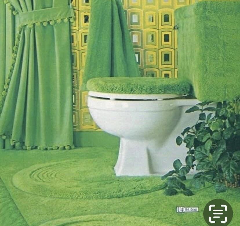 A bathroom decorated in bright green shag carpeting, covering the floor, toilet seat, tank, and lid. There is a leafy plant by the toilet and a yellow patterned wallpaper on the wall.