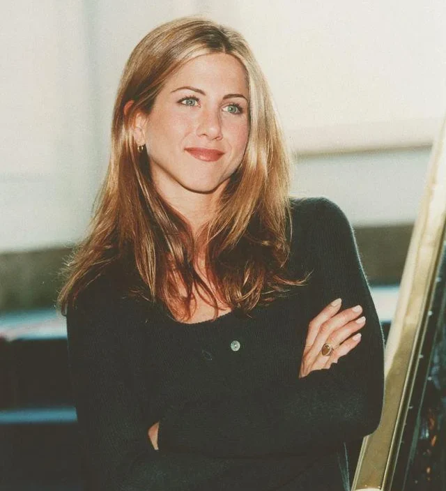 A woman with long, light brown hair stands indoors, smiling softly with her arms crossed. She is wearing a dark buttoned sweater and hoop earrings, standing near a staircase with a bright background.