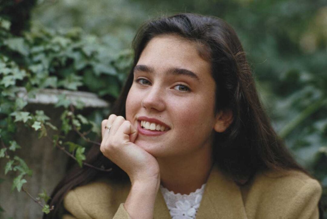 A young woman with long dark hair smiles at the camera, resting her chin on her hand. She is wearing a tan jacket and white blouse, with green foliage and a stone surface in the background.