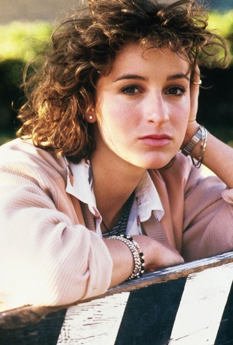 A young woman with curly brown hair rests her head on her hand, looking thoughtfully at the camera. She wears a light jacket, a patterned shirt, and silver jewelry, sitting outdoors near a striped barrier.