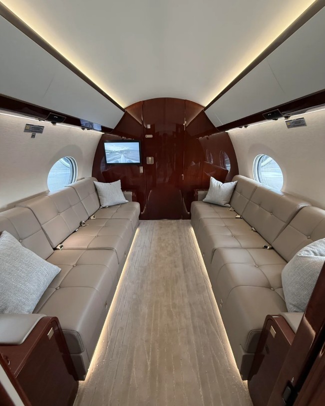 Interior of a private jet featuring two long beige leather sofas facing each other, light carpeting, round windows on each side, wall-mounted screens, and decorative pillows, creating a luxurious and modern atmosphere.