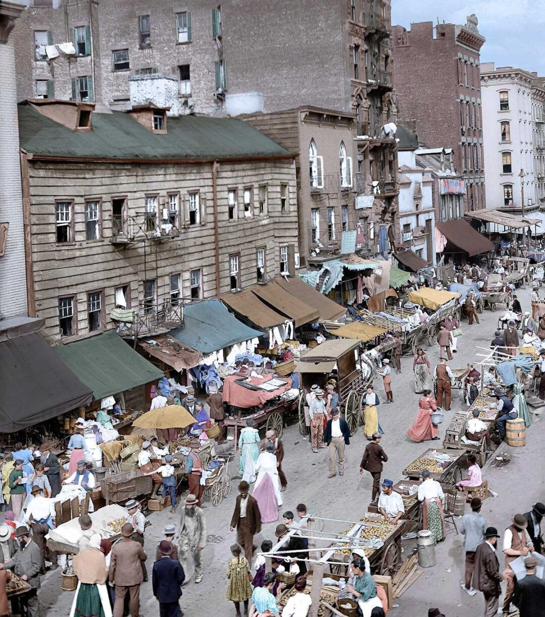 A bustling outdoor market scene in a city, with many people walking among vendor stalls selling goods; old multi-story buildings line the street in the background. The atmosphere is lively and crowded.