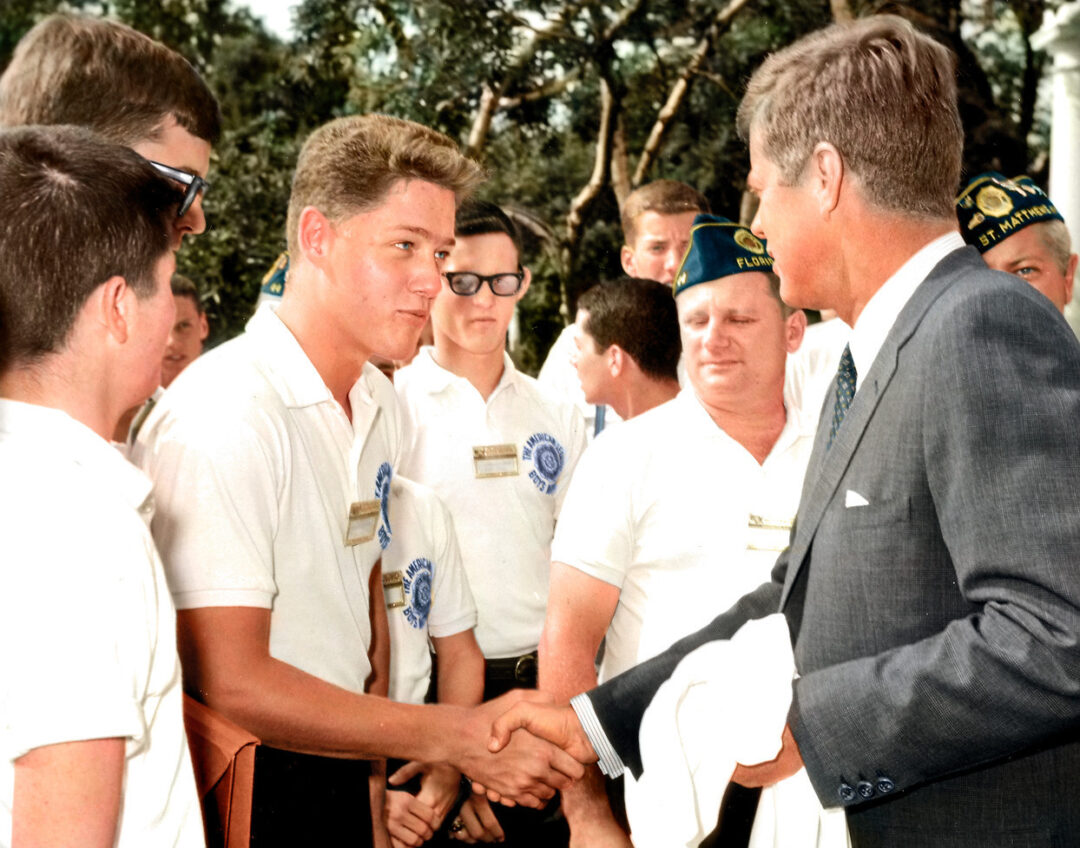 A group of young men in white shirts stand outdoors as one shakes hands with an older man in a suit. The group appears to be participating in a formal or ceremonial event. Trees and more people are visible in the background.