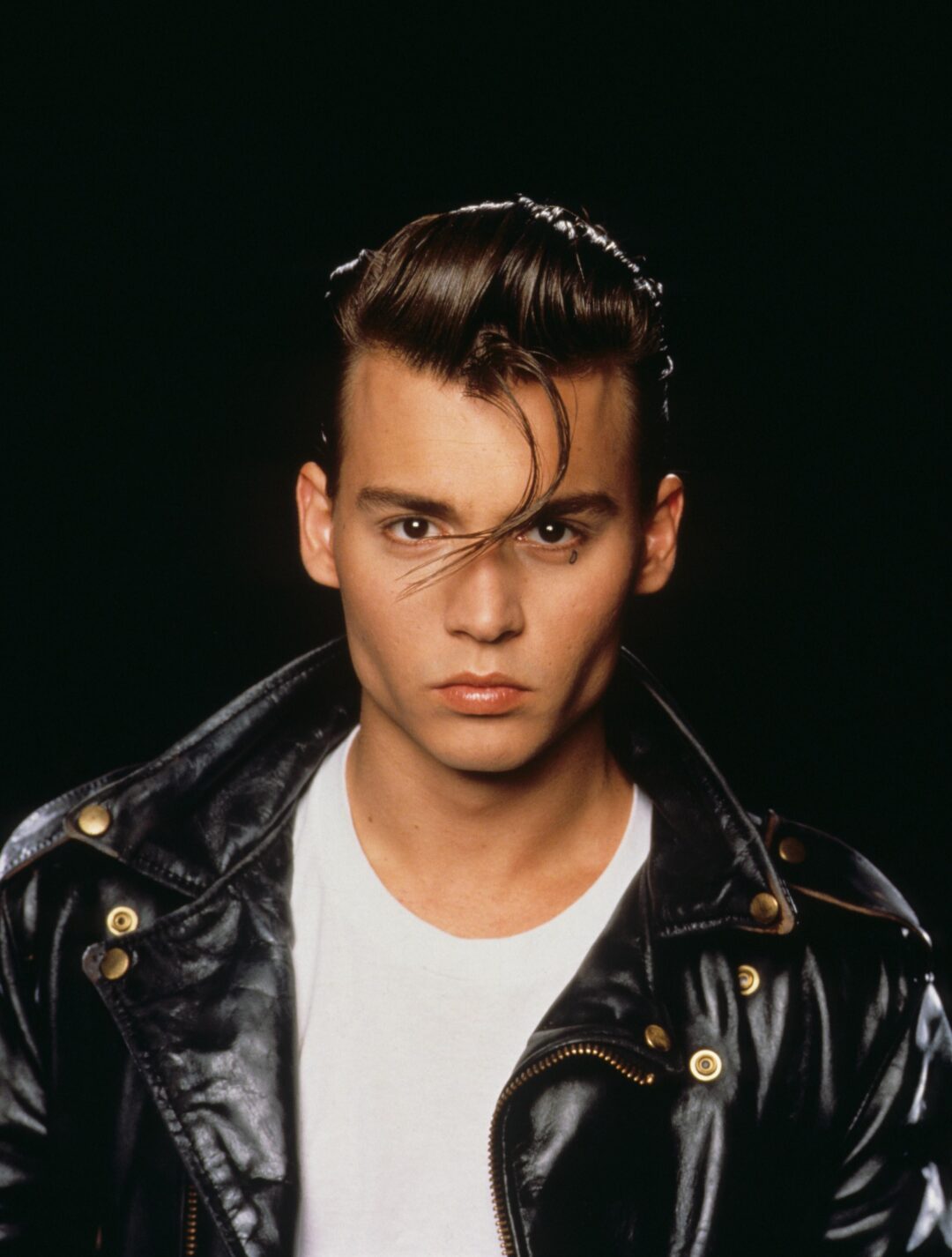 A young man with slicked-back hair and a wavy strand hanging over his forehead wears a black leather jacket and white T-shirt, posing against a dark background with a serious expression.