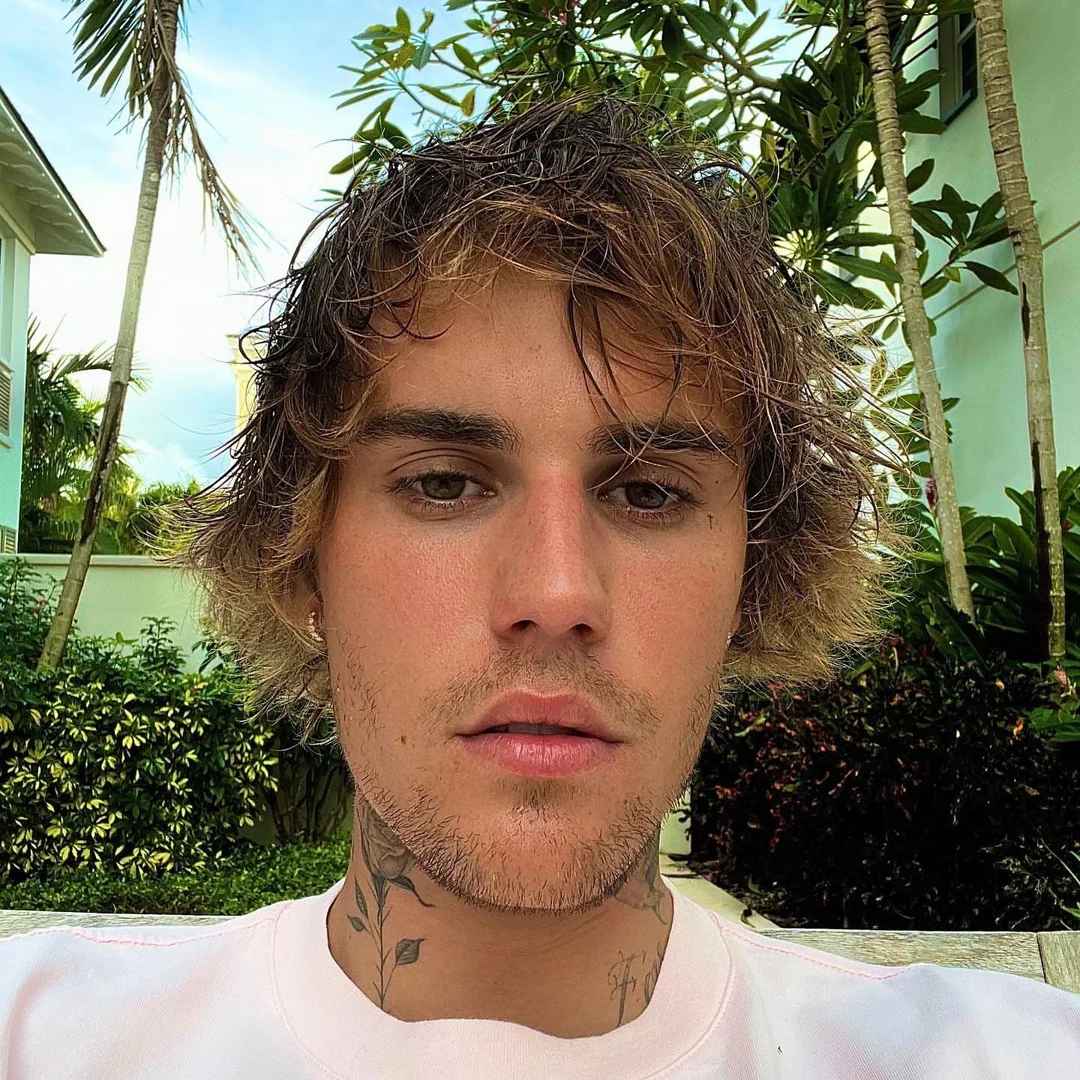 A young man with tousled brown hair and facial stubble looks directly at the camera. He has neck tattoos and wears a light-colored shirt. Lush tropical plants and buildings are visible in the background.