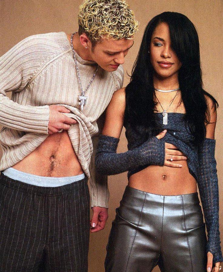 A man and a woman stand side by side, both showing their midriffs. The man lifts his sweater to reveal his stomach, while the woman places a hand on her bare stomach and looks at him. Both wear stylish, casual outfits.