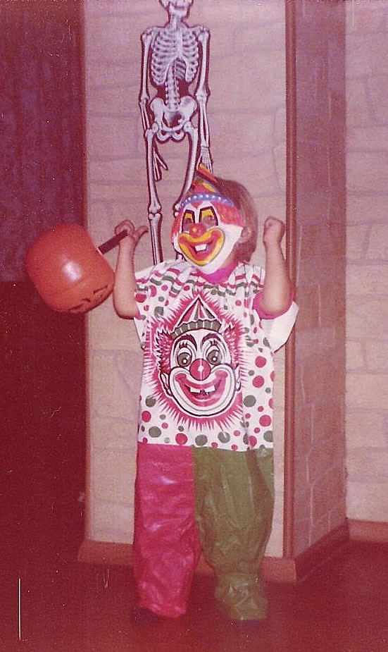 A child dressed in a colorful clown costume and mask holds a pumpkin-shaped bucket, standing in front of a cardboard skeleton decoration on a wall.