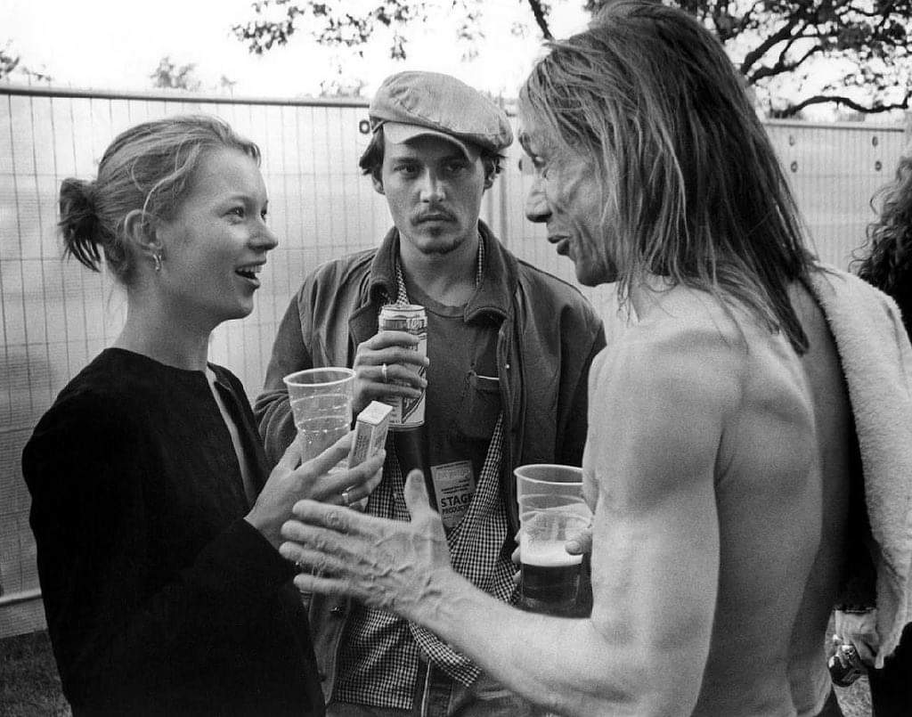 Three people talking outdoors. A woman holds a drink and smiles at a shirtless man with a towel over his shoulder. A man in the middle, wearing a cap and holding a can, looks at the camera. Trees and a fence are in the background.