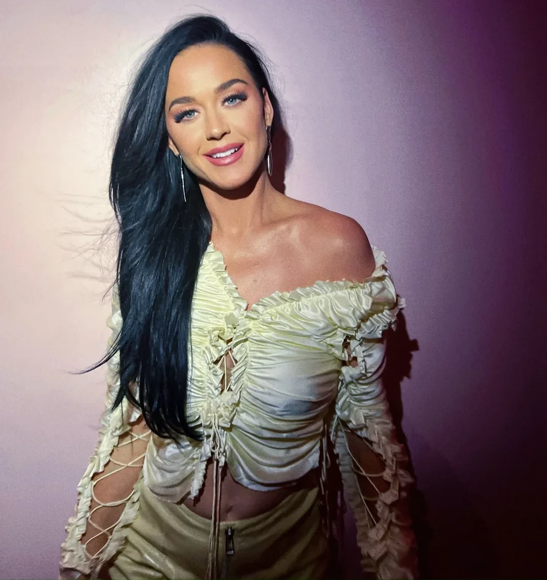 A woman with long dark hair smiles at the camera. She is wearing a pale off-the-shoulder, ruched top with lace-up sleeves and standing against a light pink background.