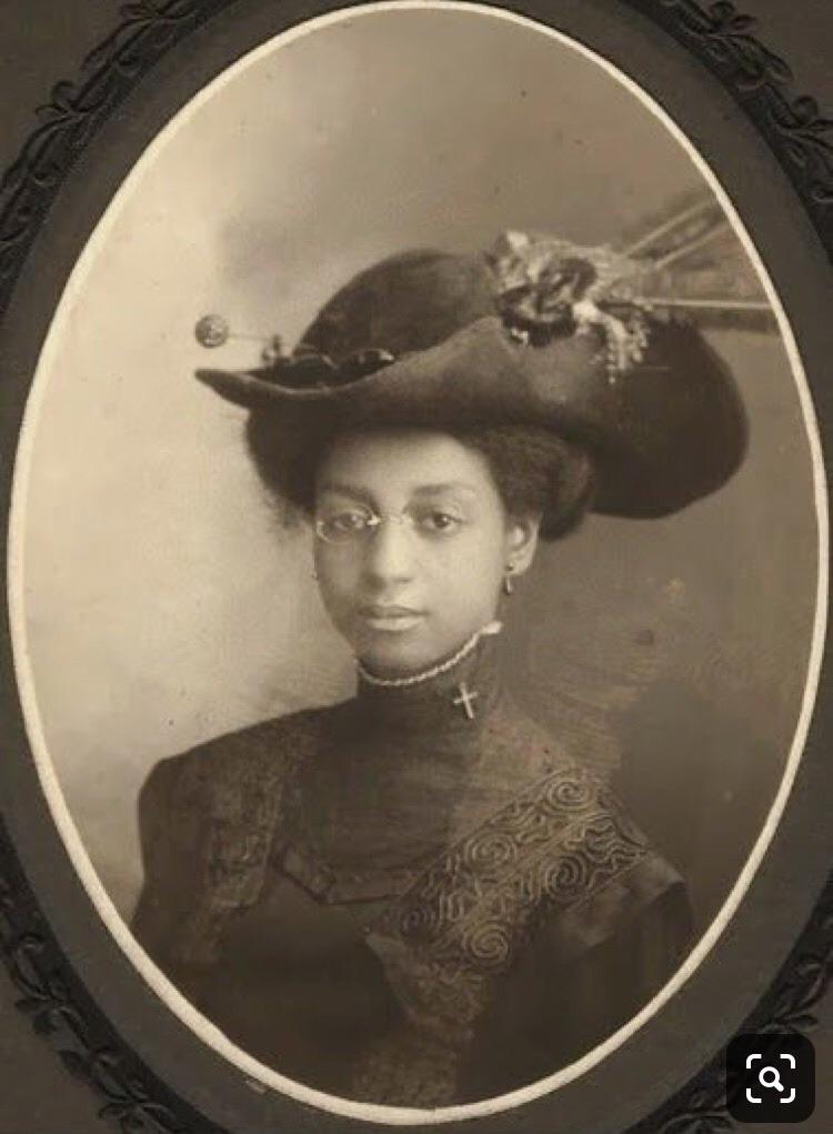 A vintage black-and-white portrait of a young Black woman wearing glasses, a high-necked dress with embroidery, a cross necklace, and an elaborate hat adorned with feathers and decorations, posed against a plain backdrop.