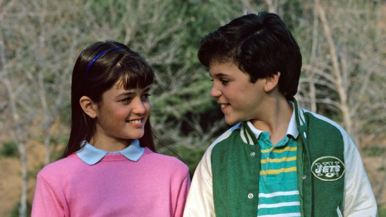 A young girl in a pink sweater and a blue headband smiles at a boy in a green varsity jacket and striped shirt. They are standing outdoors in front of trees, looking at each other warmly.