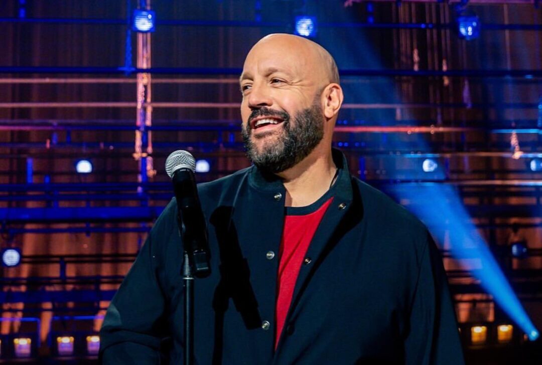 A smiling, bald man with a beard stands at a microphone on a stage, wearing a dark jacket over a red shirt. Blue and purple stage lights shine in the background.