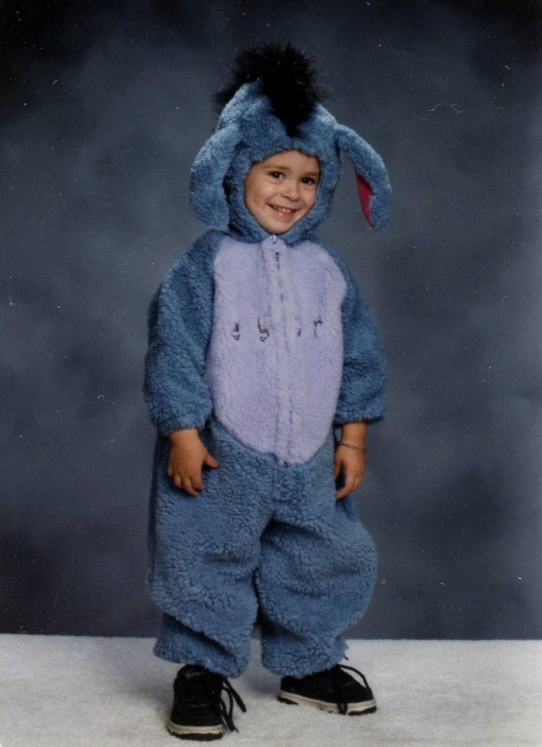 A young child smiles while wearing a fuzzy Eeyore costume with floppy ears, a tail, and black sneakers, standing against a gray studio backdrop.