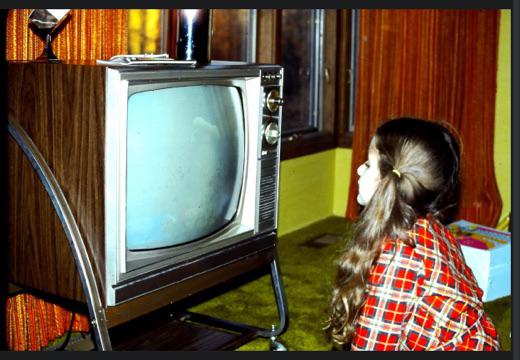 A young girl in a red plaid shirt sits on green carpet, watching a vintage box television in a room with orange curtains and wood-paneled walls.