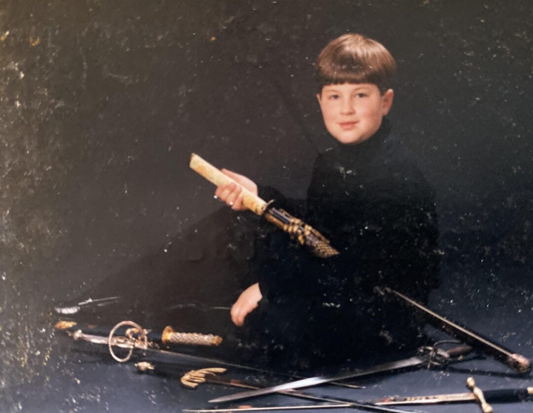 A young boy with short light brown hair, dressed in black, sits on the floor against a dark background, holding a decorative sword. Several other swords and ornate blades are arranged on the ground in front of him.