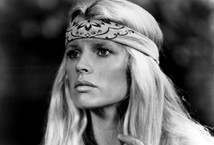 A black and white photo of a woman with long, blonde hair wearing a patterned headband across her forehead, looking serious and slightly to the side.