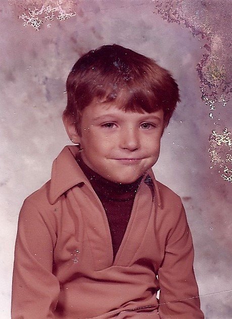 A young boy with short brown hair sits for a school portrait, wearing a tan collared shirt over a dark turtleneck, smiling slightly against a neutral mottled background.