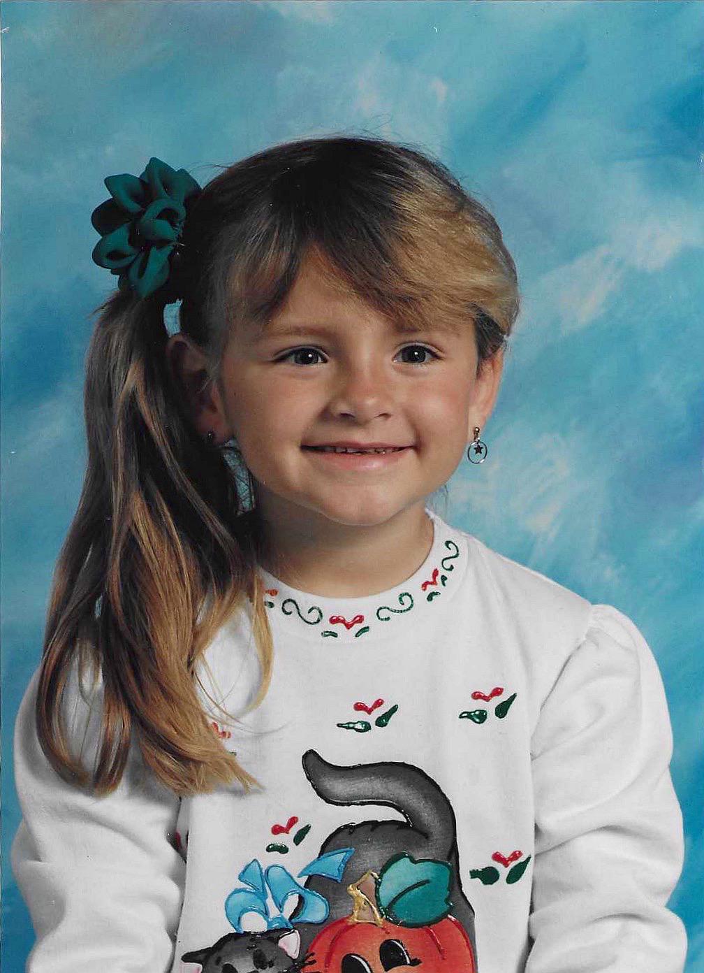 A young girl with long blond hair in a side ponytail and a green hair bow smiles at the camera. She wears a white shirt with a cat and pumpkin design, and sits in front of a blue studio background.