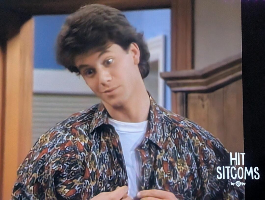 A young man with dark hair, wearing a colorful patterned shirt over a white t-shirt, stands indoors with a slightly confused or surprised expression. “HIT SITCOMS” is displayed in the lower right corner.